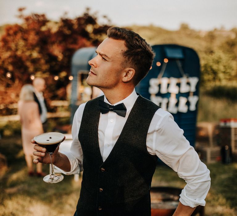 Groom in white shirt, waistcoat and bow tie looks off into the distance with hand on hip whilst holding an espresso martini during golden hour at garden wedding reception