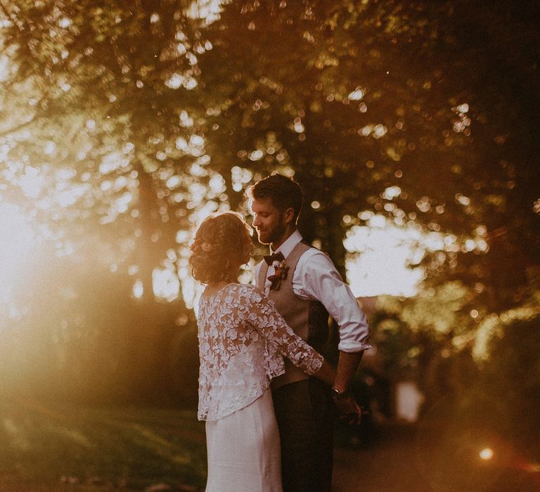 Bride in white Rime Arodaky wedding dress with mesh floral embroidered top stands with arms around groom in white shirt and light brown waistcoat under trees during golden hour at garden wedding with burnt orange wedding theme