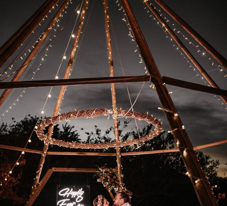 Bride in a lace wedding dress and groom in a white shirt and linen trousers dancing on wool rugs under a naked tipi decorated in fairy lights 