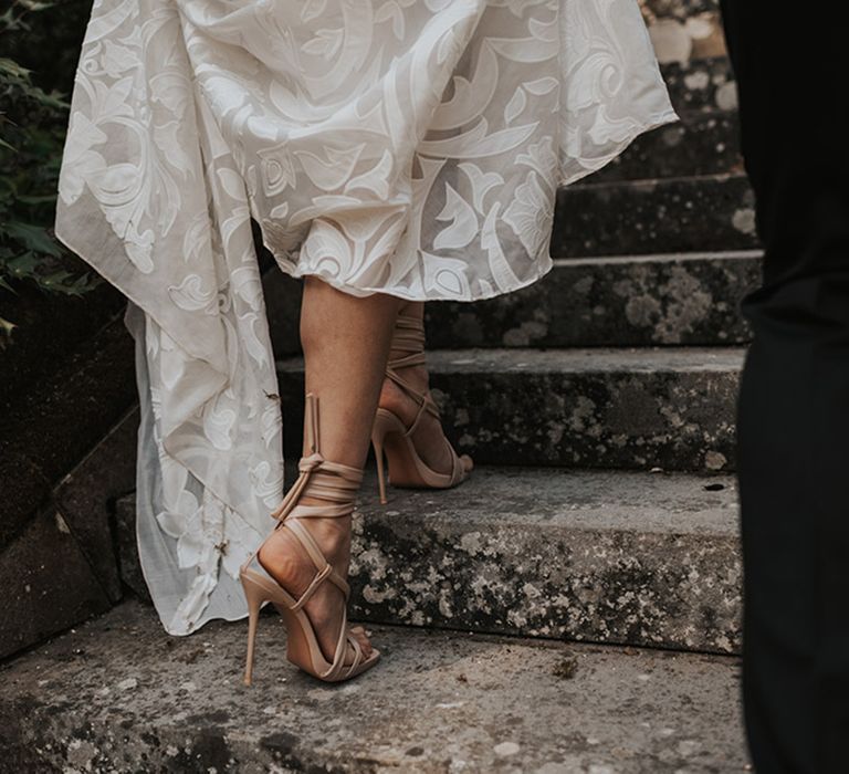 Bride in beige ankle strap sandals walking up the steps holding her wedding dress train 