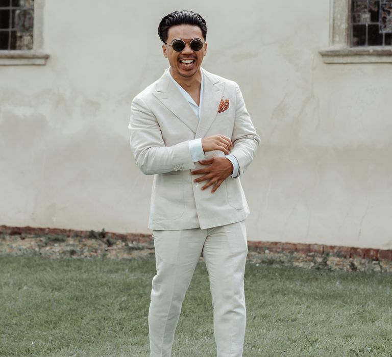 Groom in round sunglasses, shawl collared shirt and linen suit laughs whilst standing outside church before Wasing Park wedding