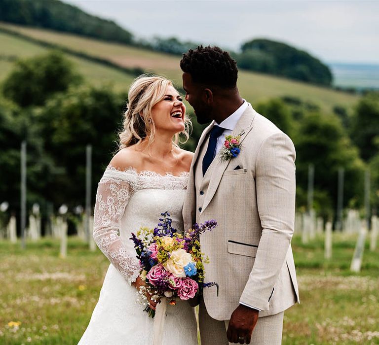 Groom in a beige suit with navy check detail smiling at his bride in a lace Bardot wedding dress holding a colourful wedding bouquet 