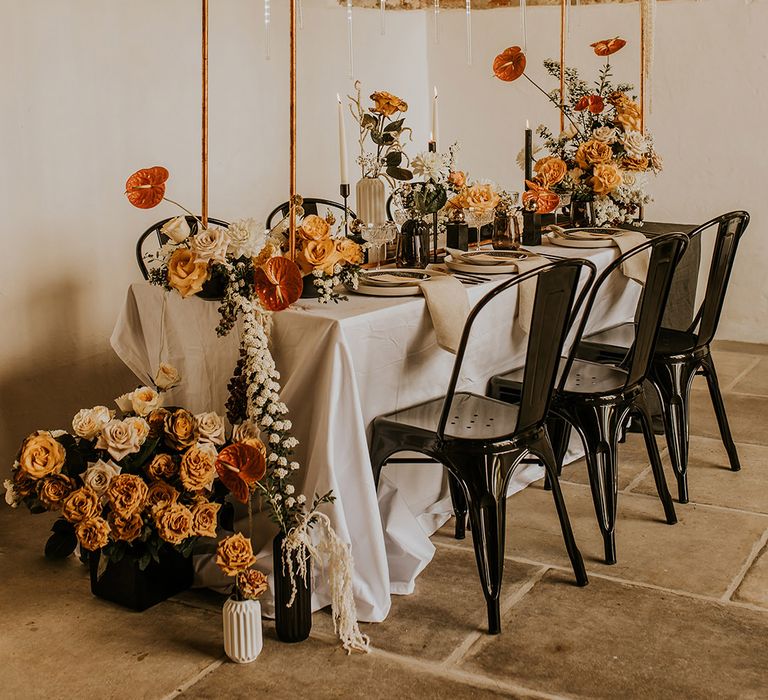 Stylish wedding reception table decor with black chairs, copper installation frame and orange and yellow wedding flowers 