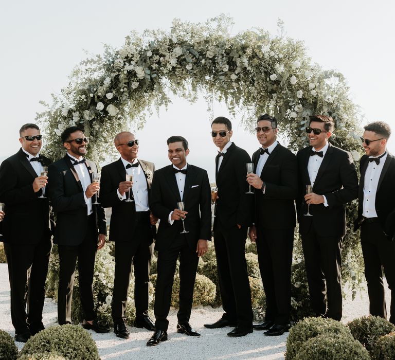 Groom and his groomsmen wear black tie in front of floral installation in Greece  | Hannah MacGregor Photo & Film