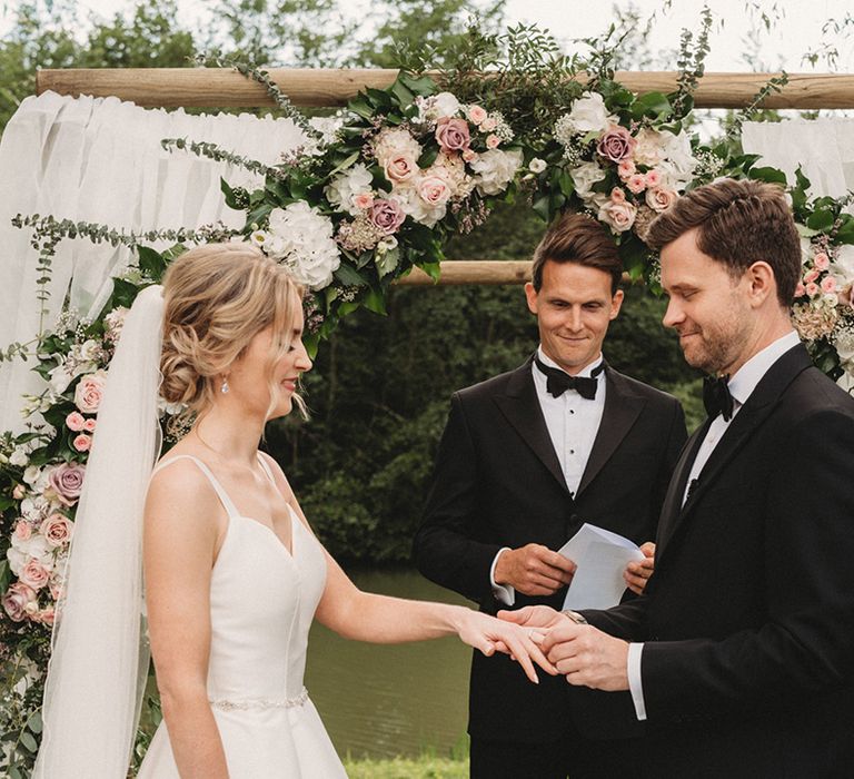 Outdoor wedding ceremony with pink and white floral arch at Chateau Lagorce with groom in a black tuxedo putting a ring on his brides finger