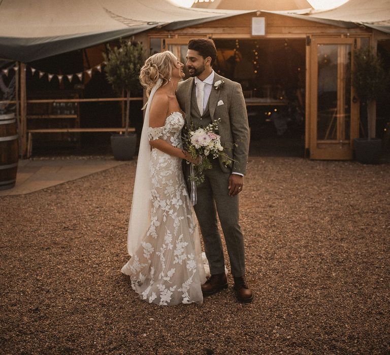 Bride in white off the shoulder Enzoani wedding dress and veil holding mixed bridal bouquet hugs groom in grey three piece suit as they stand outside of tipi wedding venue during Inkersall Grange Farm wedding