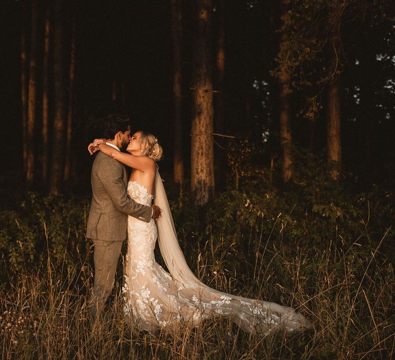 Bride in off the shoulder Enzoani wedding dress and veils kisses groom in grey suit during golden hour in woodland at Inkersall Grange Farm wedding