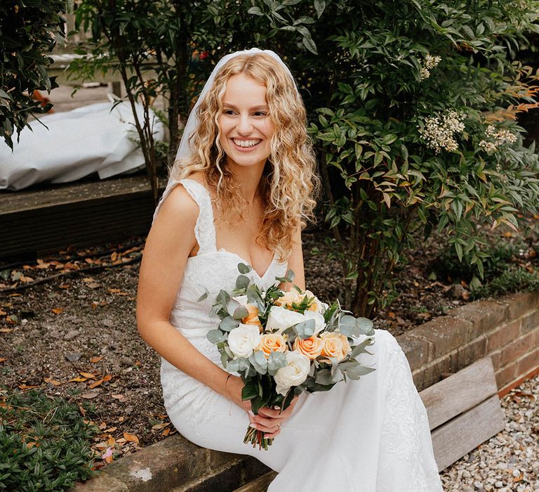 Bride sits down outdoors whilst holding her pastel peach bouquet and wearing Wed2B wedding gown with added lace straps