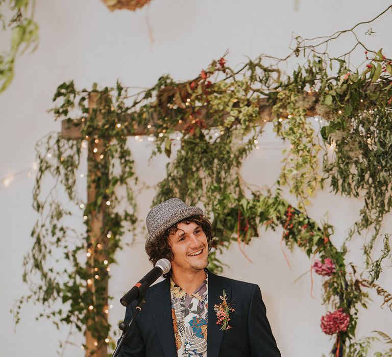 Groom stands in front of green DIY foliage on his wedding day