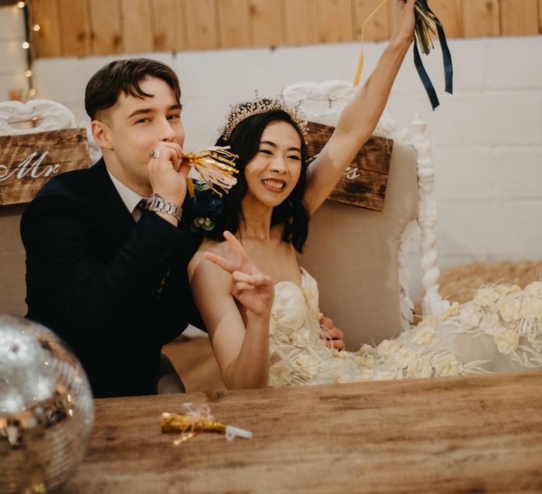 Bride in strapless feathered wedding dress holds up peace sign and tropical wedding bouquet as groom blows party blower with streamers at rustic wedding reception