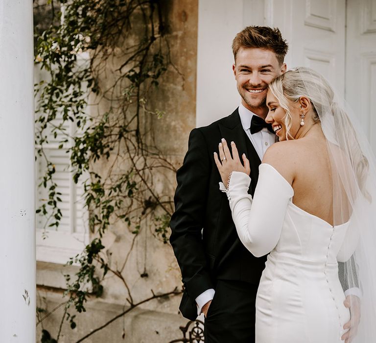 Bride in a fitted strapless wedding dress with buttons down the back embracing her husband in a tuxedo on the grounds at Northbrook Park 