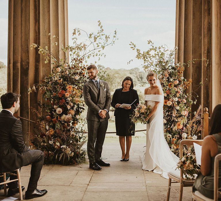 Bride & groom stand in front of floral archway on their wedding day during ceremony