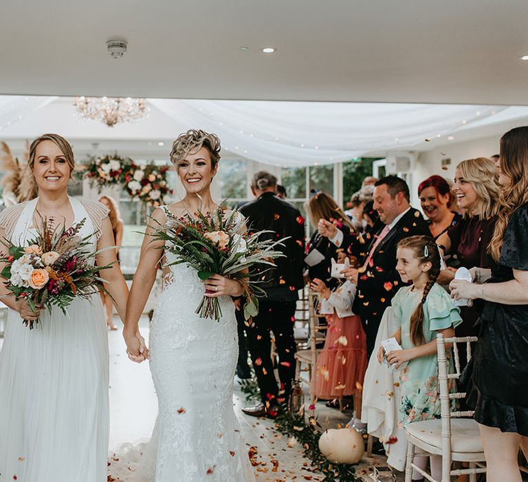 Brides hold hands on their wedding day as they walk down the aisle