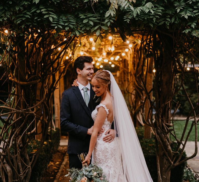 Bride in open back lace and tulle wedding dress and veil stands hugging groom in dark suit in the grounds of The Tythe Barn in Launton