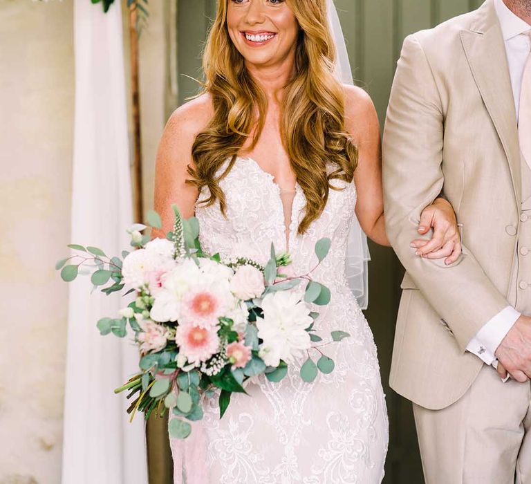 Bride walks down the aisle wearing her hair long and slightly curled whilst holding floral bouquet