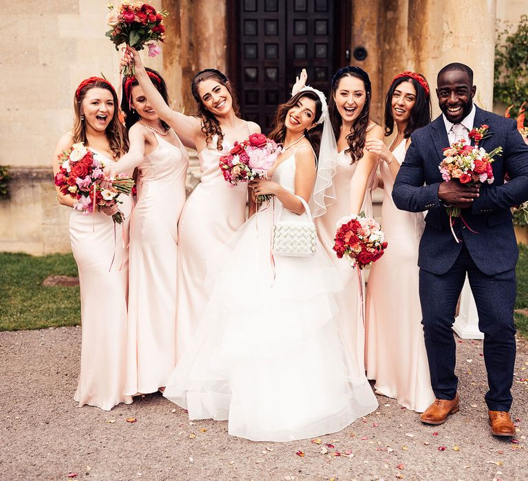 Bride in a line tulle skirt wedding dress holding beaded bag and pink, white and red wedding flowers stands with bridesmaids in pink satin bridesmaid dresses and bridesmaid in blue suit as they wave bouquets