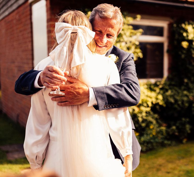 Bride in wedding dress and satin hair bow hugs man in blue suit holding glass of champagne in garden