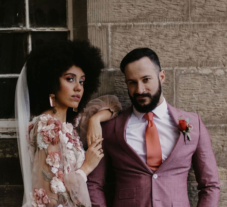 Black bride with afro hair in a 3D flower appliqué wedding dress with her groom in a salmon coloured wedding suit 