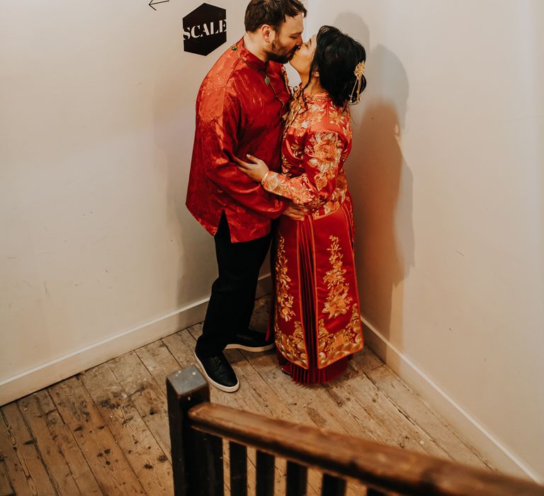 Bride in red and gold traditional Chinese tea ceremony dress kisses groom in black trousers and red traditional Chinese clothing