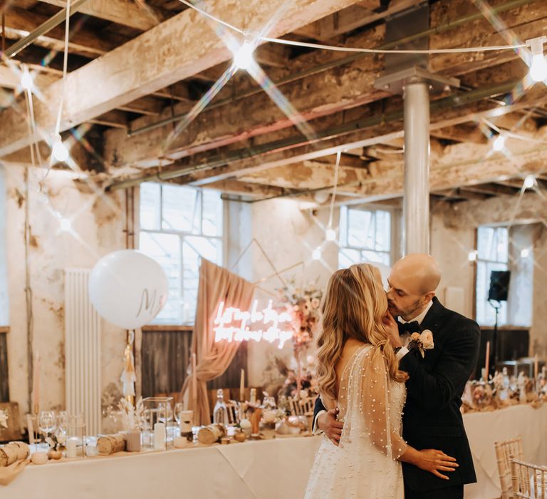 Bride in white wedding dress with train and pearl details kisses groom in navy suit as they stand in Holmes Mill wedding reception room with neon wedding sign