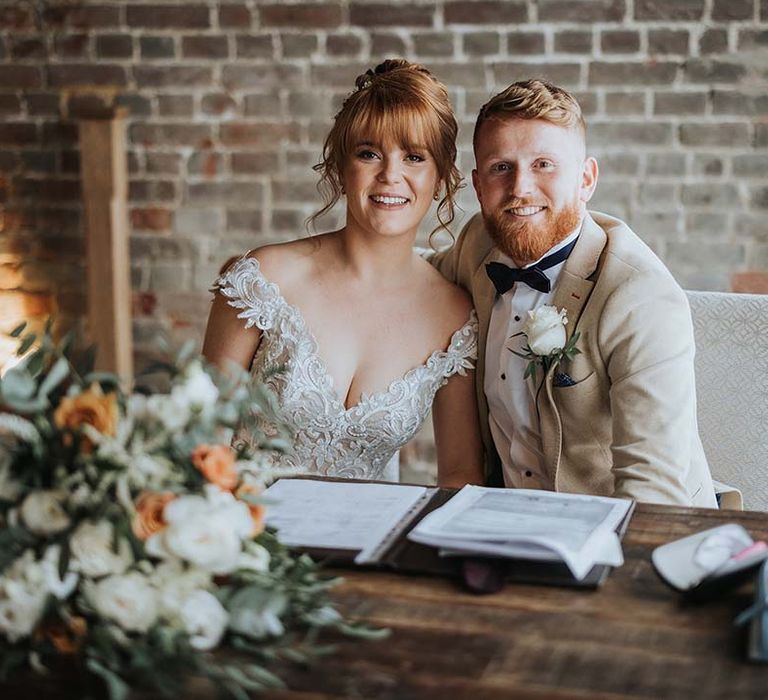 Bride and groom smile together as they sign the register with white and orange colour scheme for spring wedding