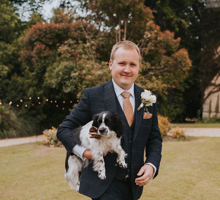 Groom in checkered suit and snake print tie holds his pet dog as he walks along the grounds of the wedding venue