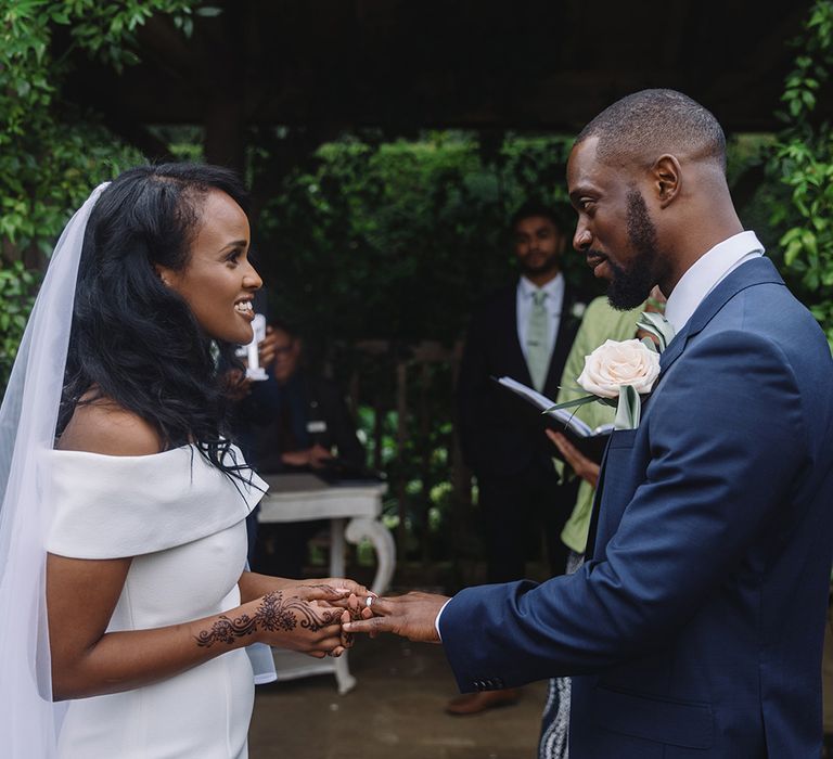 Bride in off the shoulder wedding dress places ring on the groom's finger who wears a white rose boutonnière