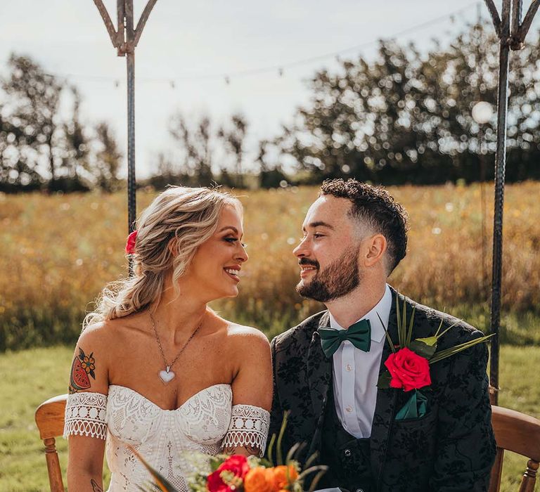 Groom in textured tuxedo with dark green bow tie and red rose buttonhole with bride in boho lace wedding dress and colourful bouquet 