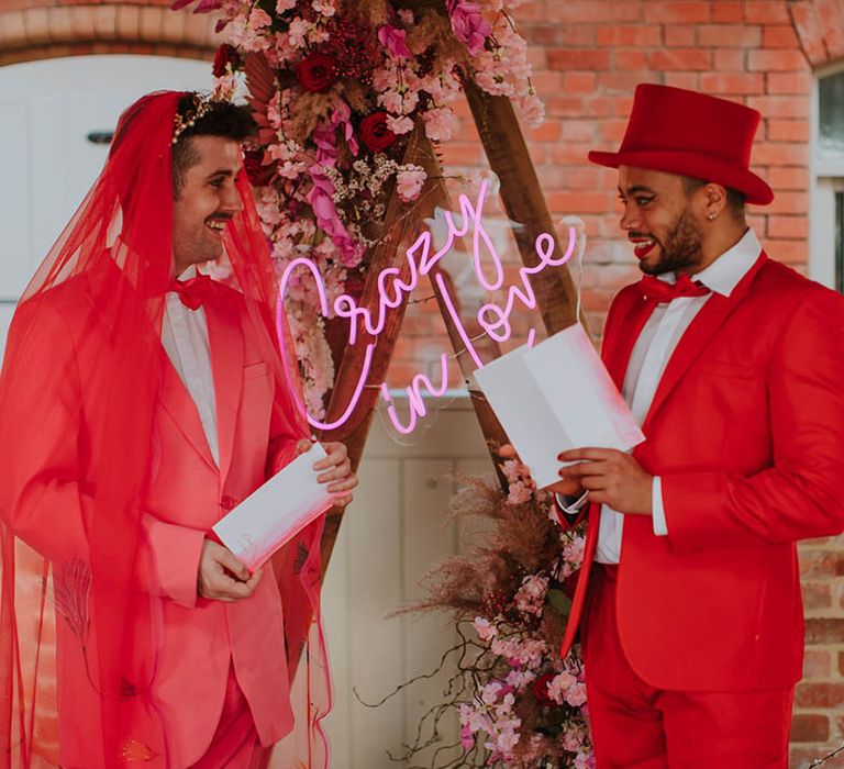 LGBTQI+ couple exchanging vows at Prestwold Hall Barns in a pink suit with red veil and a red suit and top hat 