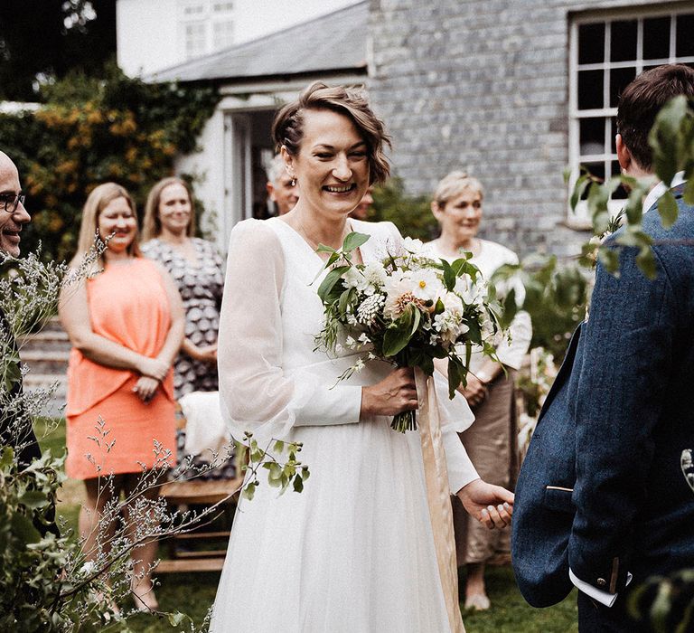 Bride smiles at the altar for outdoor wedding ceremony at Treseren holding white flower bouquet including cosmos and dahlias