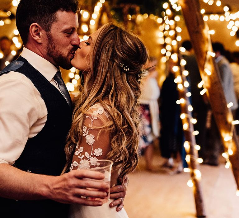 Bride and groom kiss in front of fairy light as the groom holds a beer 