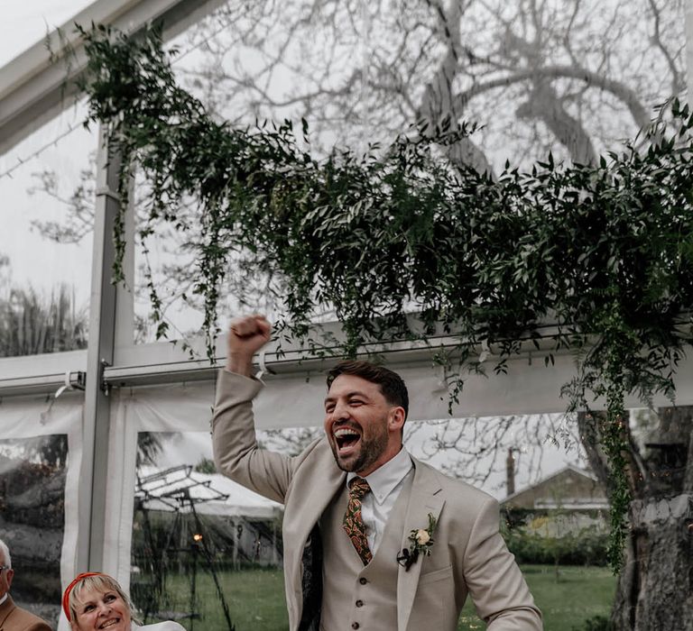 Groom in a stone coloured three-piece with paisley tie fist bumping he air during the clear marquee wedding reception  