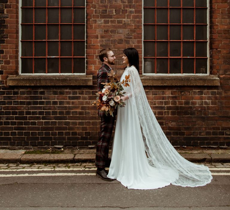 Bride wears lace cape on her wedding day as she stands in front of brick wall with her groom 