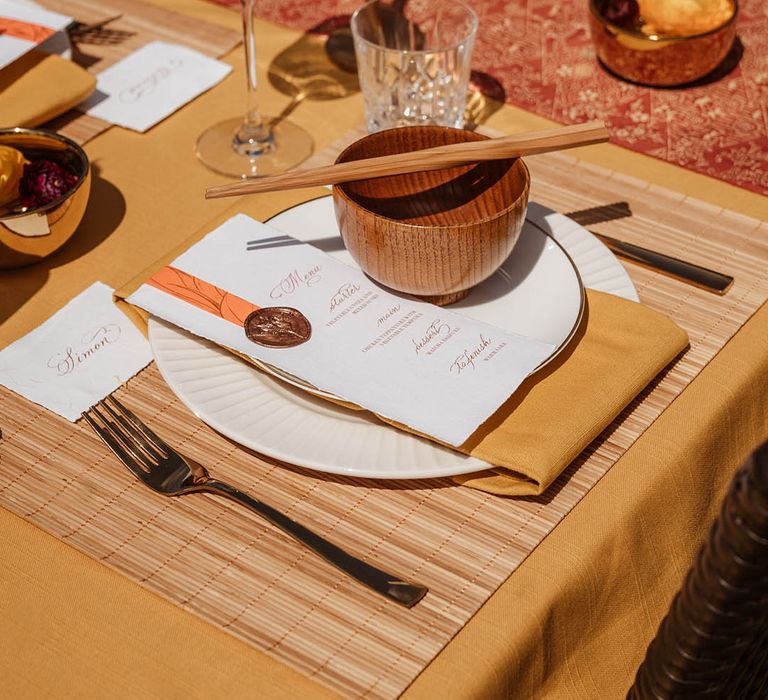 Tablescape with yellow table rubber, bamboo place mat, gold cutlery, brown rice bowls, wooden chopsticks, gold candlesticks and menus card for Japanese wedding 