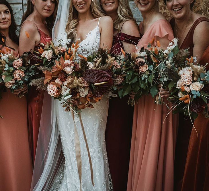 Bride stands with her bridesmaids wearing different styled bridesmaid dresses in differing shades of orange and red