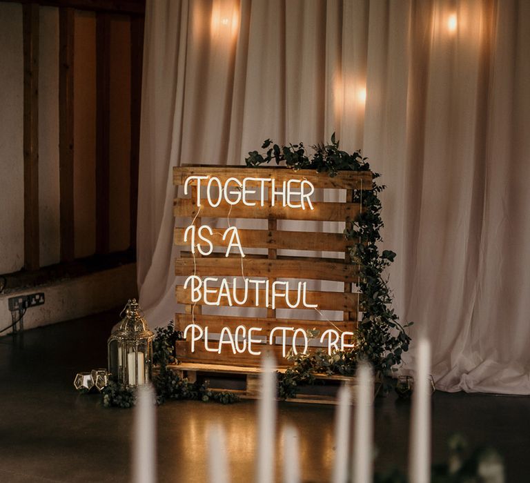 Wooden pallet decorated with foliage and candle lanterns with white neon sign reading 'Together Is A Beautiful Place To Be'