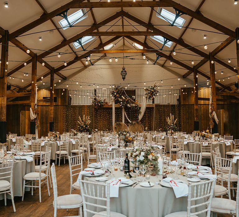 Grey wedding table settings with venue decorated with festoon lighting, pampas grass, peacock chairs and white wedding drapes 