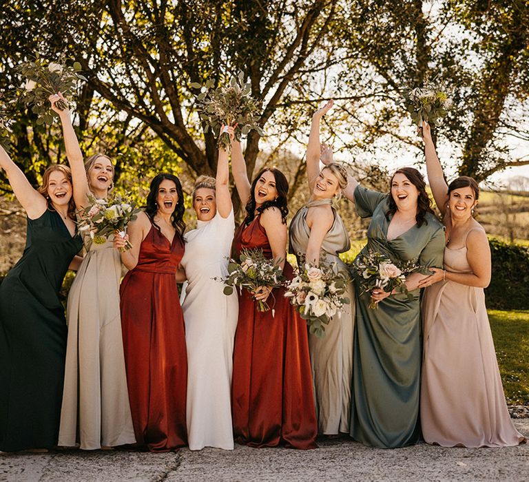 Bridesmaids in different coloured satin dresses lift their bouquet with the bride 