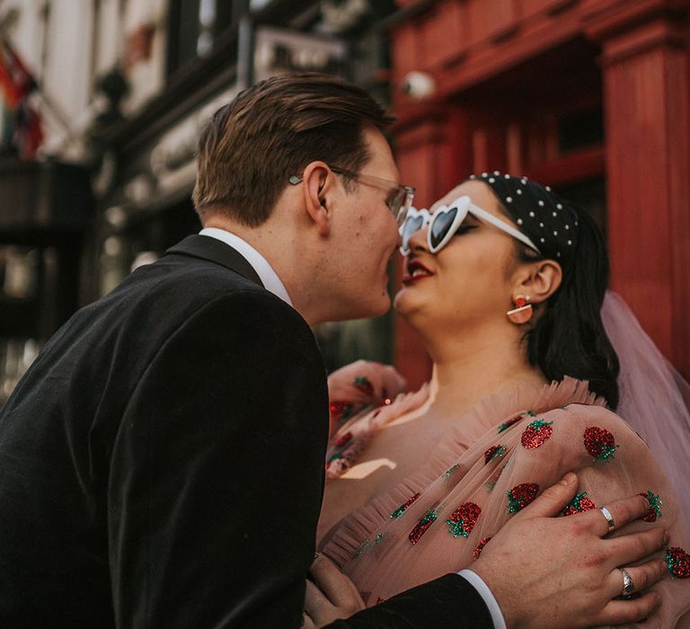 Bride wears heart-shaped sunglasses and pink chiffon dress as she kisses her groom outdoors in London on their wedding day