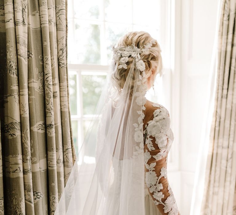 Bride stands beside window as she wears cathedral veil complete with lace edging and Pronovias wedding dress 