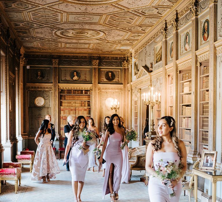 Bridesmaids wearing lavender bridesmaid dresses and holding brightly coloured floral bouquet walk through grand room at Syon Park