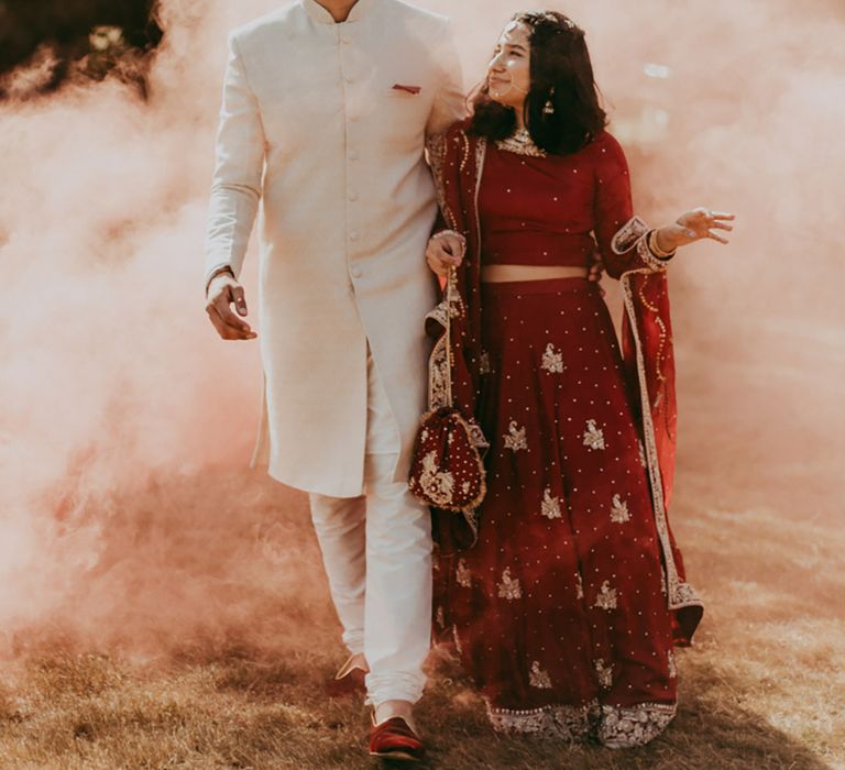 Bride in red and gold embellished bridal separate walks through orange smoke bomb with her groom