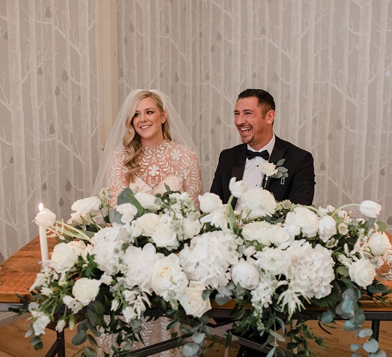 Bride & groom sit at wooden table with white florals and green foliage