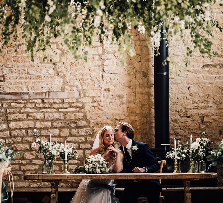 Groom kisses the bride on the cheek as they sit together to sign the register at the Old Gore Barn