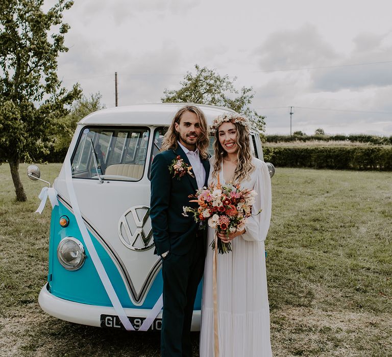 Bride & groom stand in front of VW camper van as bride holds colourful floral bouquet tied with champagne coloured ribbon