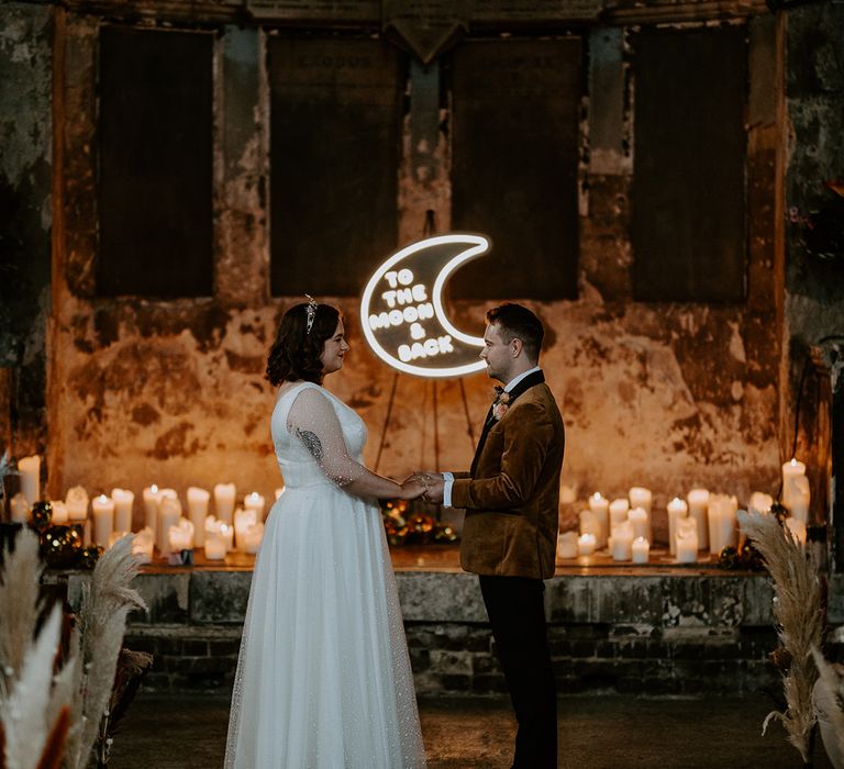 The couple look lovingly at each other surrounded by warming wedding lighting created by candles and a moon neon sign