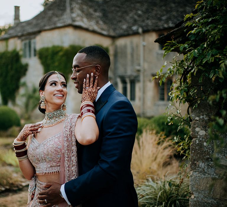 Panjabi Bride and Jamaican Groom pose in the grounds of country wedding venue wearing a golden bridal Lehnga and Navy suit