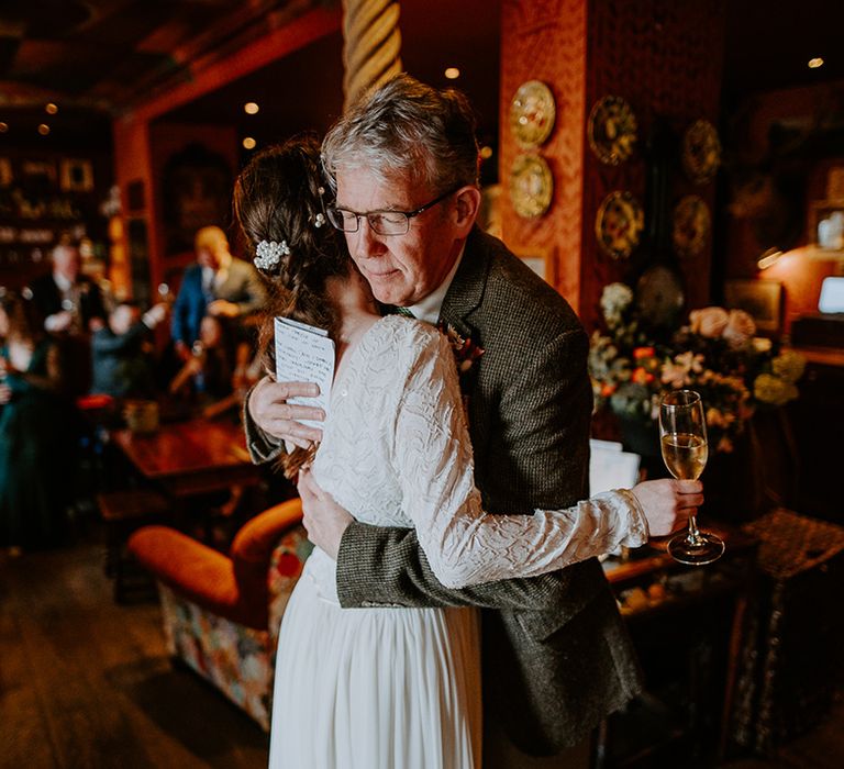 Bride holding a Champagne glass wearing long sleeve lace vintage wedding dress with drop back detail, pearl earrings and pearl hair accessories hugging her dad at intimate London wedding