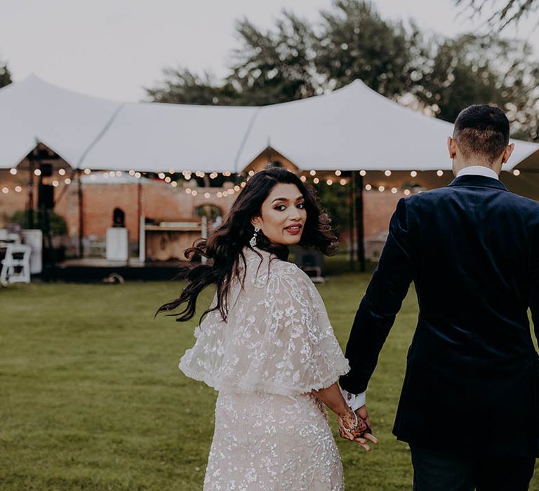 Bride wears Needle & Thread reception dress and her black hair in loose curls as she walks alongside her groom in velvet suit