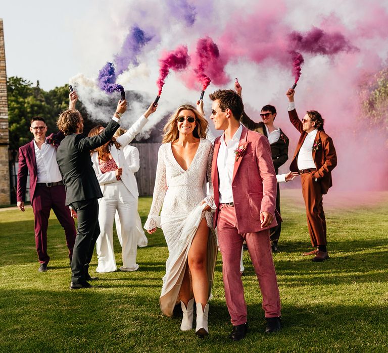 Groom in pink corduroy suit with pink and red rose, foliage and dried flower boutonniere and sunglasses holding hands with bride in v-neck long sleeve boho wedding dress with bridesmaids in bridesmaid jumpsuits and guests doing pink and purple smoke bomb photos